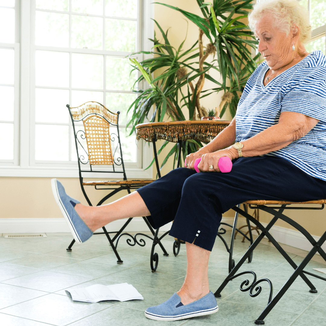 Elderly woman sitting in a chair indoors, lifting pink dumbbells with her legs, papers on the floor, and plants in the background. - Home Instead