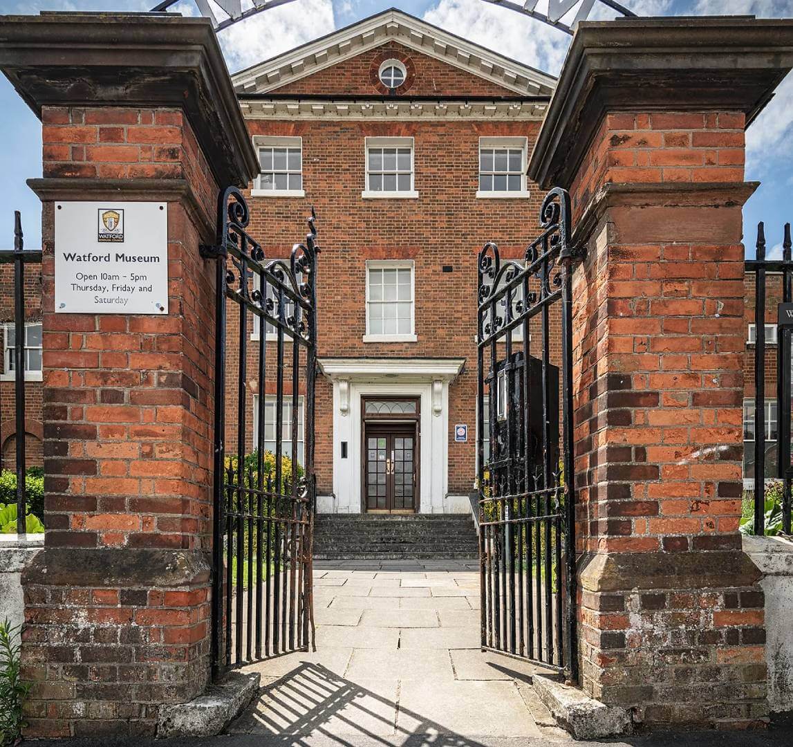 A brick building with large white-trimmed windows, iron gates open in front, and sign reading "Watford Museum. - Home Instead