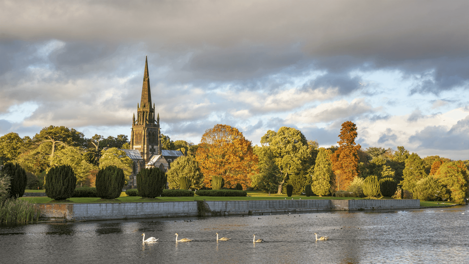Serene park with a church, autumn trees, swans swimming in a lake, and a cloudy sky. - Home Instead