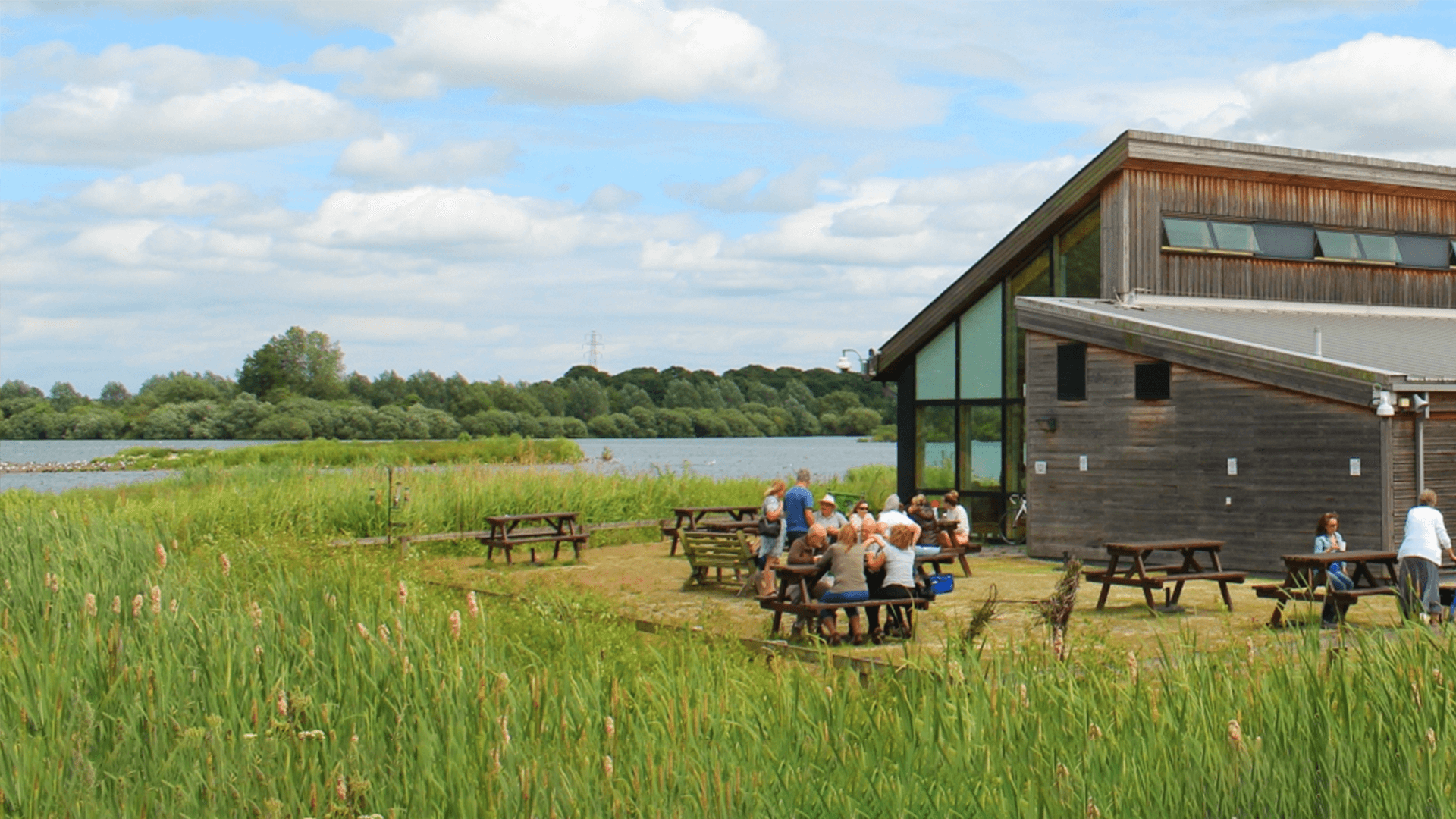 People sitting at picnic tables outside a modern wooden building near a lake with lush greenery and a blue sky. - Home Instead