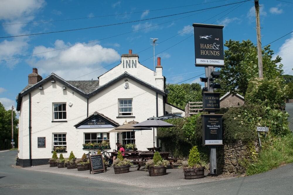 A white building with a sign reading "Hare & Hounds Inn & Dining" and outdoor seating under umbrellas on a sunny day. - Home Instead