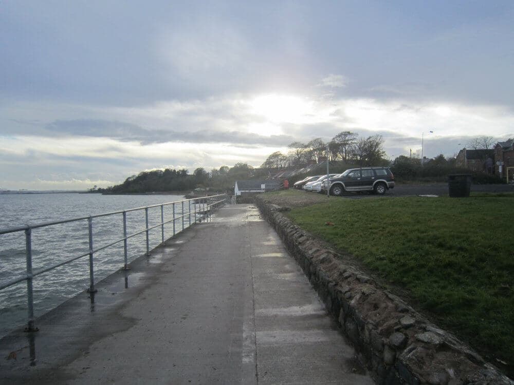 A seaside pathway with a metal railing, adjacent to parked cars and a grassy area, under a cloudy, sunlit sky. - Home Instead