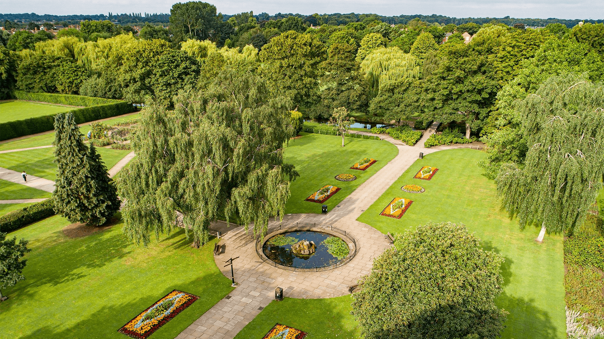 Aerial view of a landscaped park with flower beds, trees, pathways, and a circular pond at the center. - Home Instead