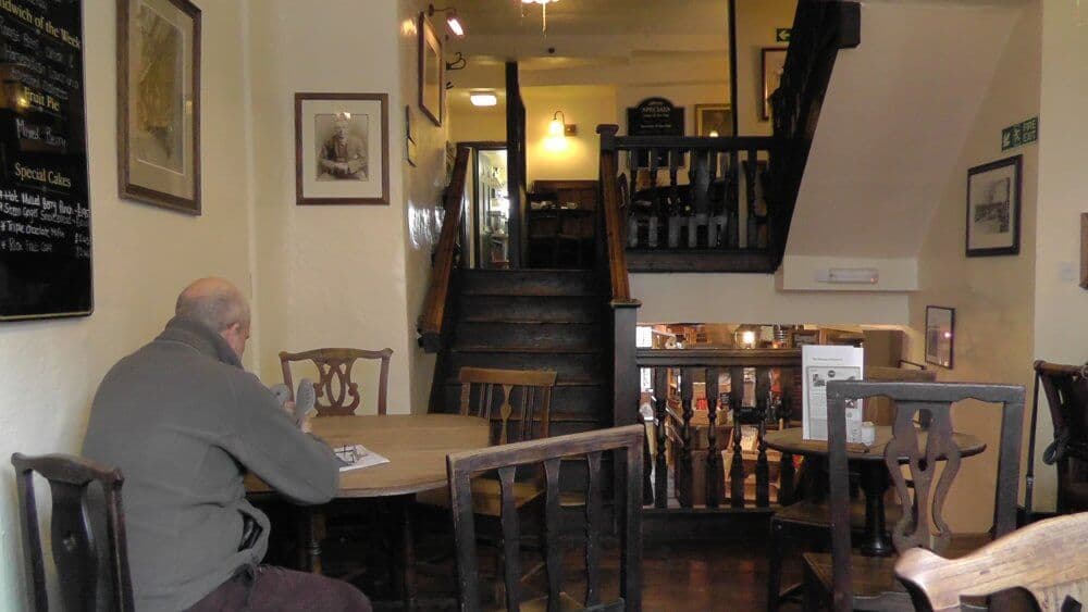 A person sits alone at a wooden table in a cozy, dimly lit café with wooden furniture and a staircase in the background. - Home Instead