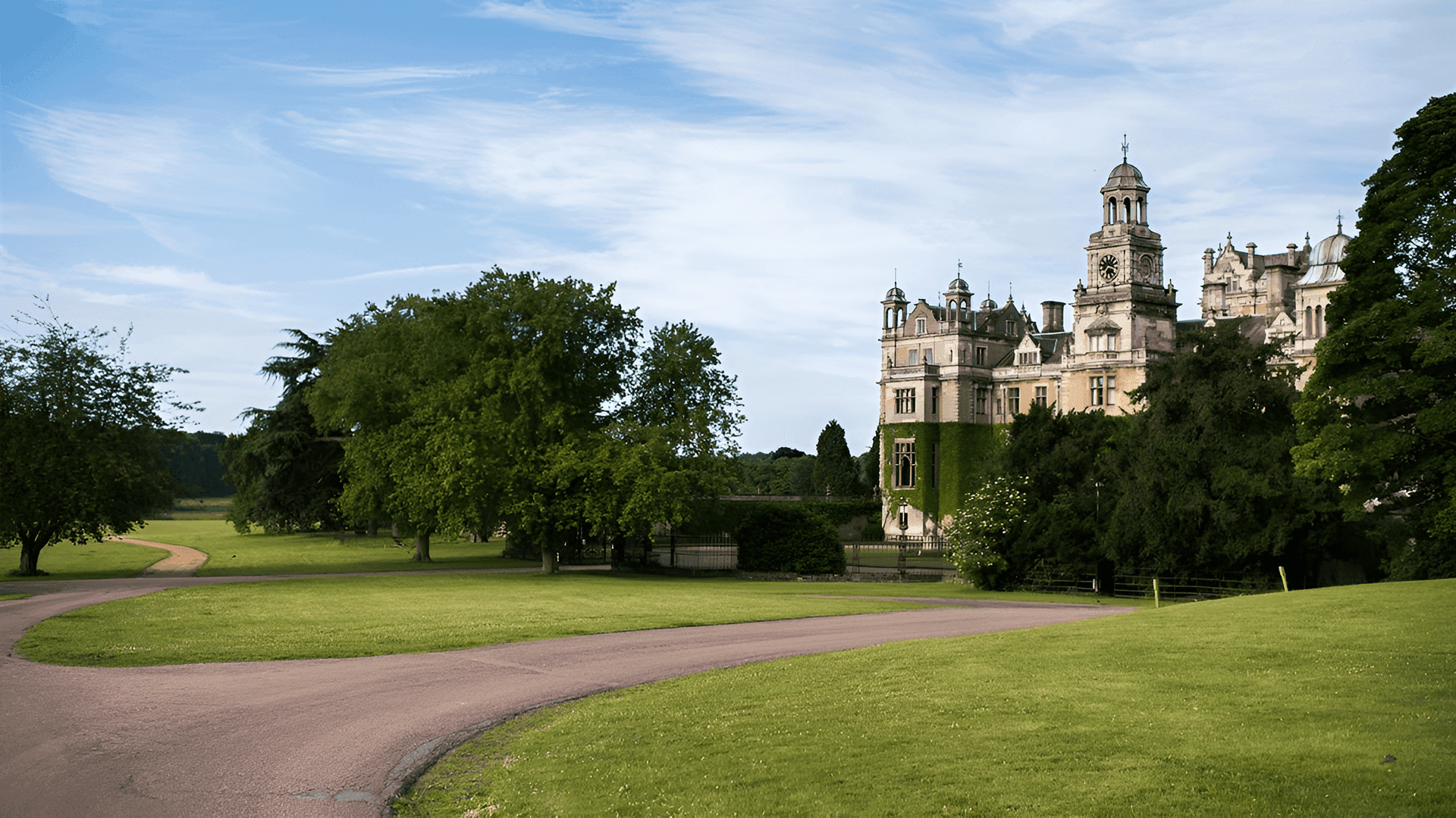 Large historic mansion with tall towers surrounded by lush green lawns and trees under a blue sky with clouds. - Home Instead