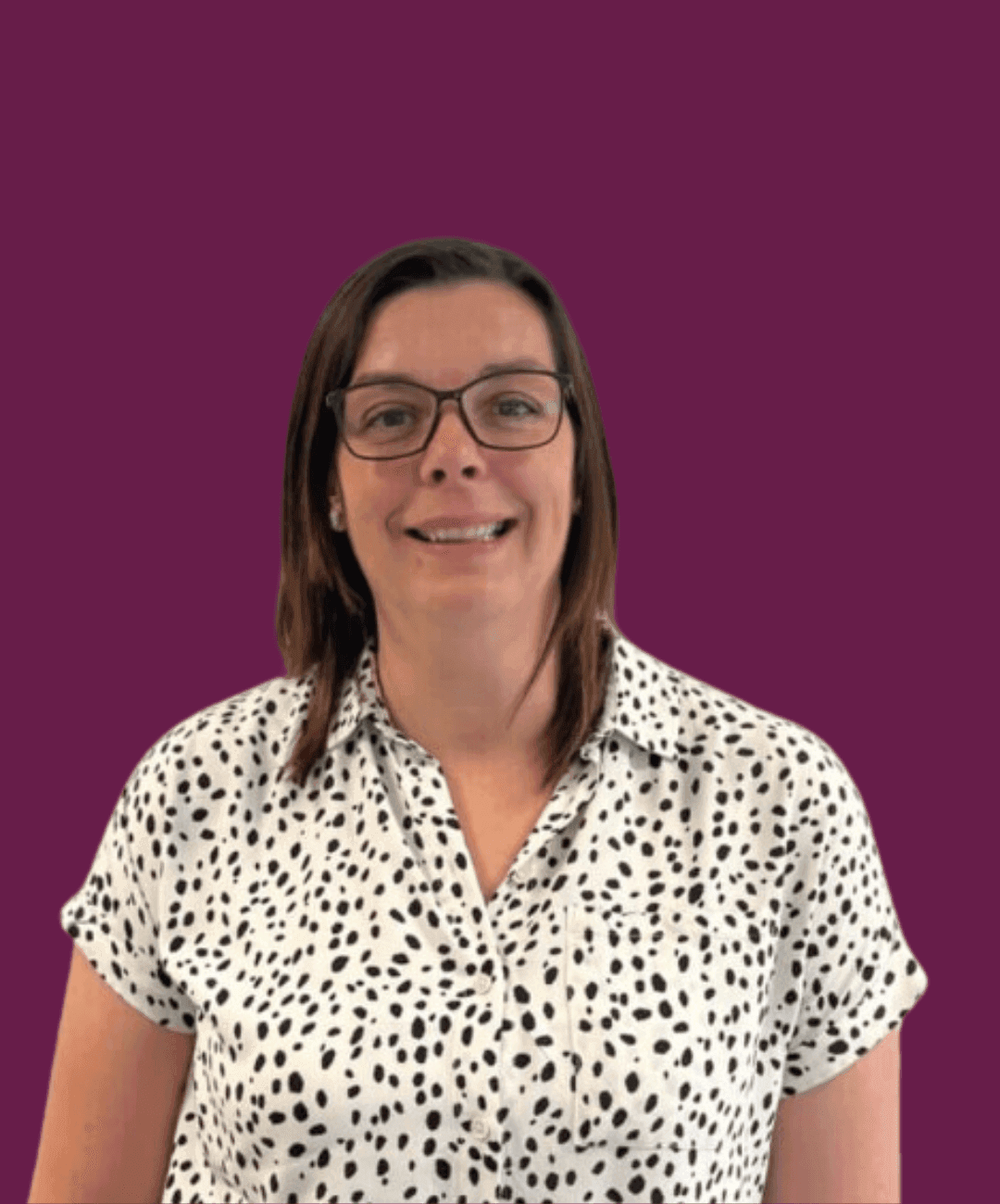 A woman with brown hair and glasses smiles against a solid maroon background, wearing a white polka-dot shirt. - Home Instead