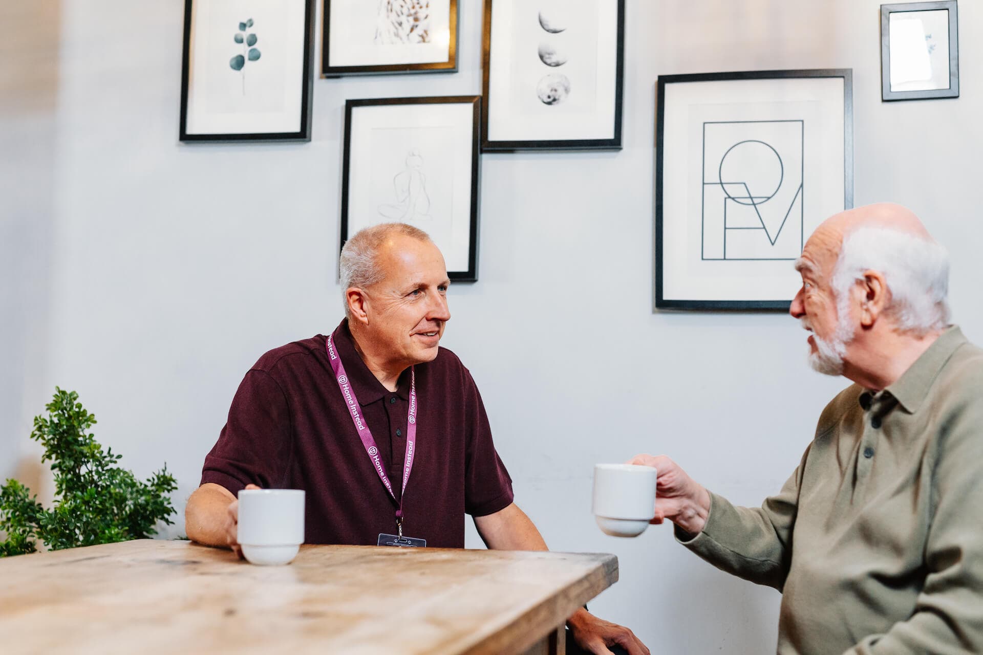 Two older men enjoy a conversation with coffee at a wooden table, with framed art hanging on the wall in the background. - Home Instead