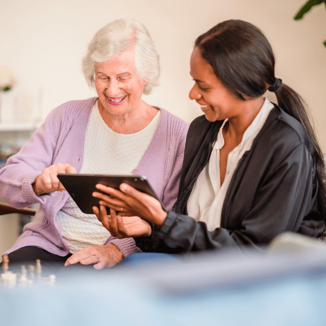 An elderly woman and a younger woman smile while looking at a tablet together in a cozy room. - Home Instead