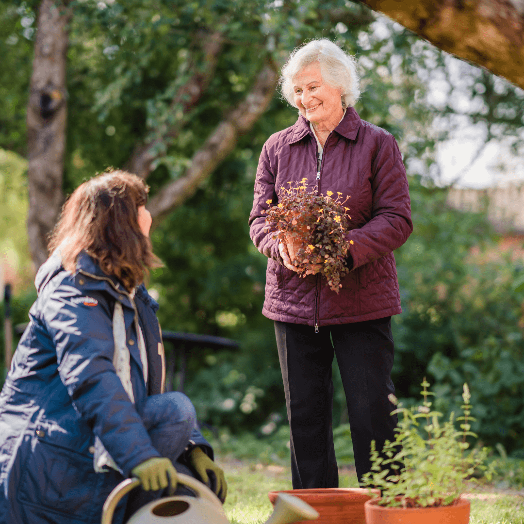 Two women enjoying gardening outdoors, one holding a plant while the other kneels beside pots and a watering can. - Home Instead