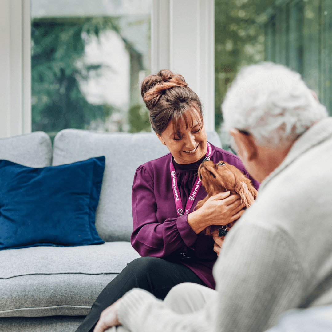 A caregiver and an elderly man sit on a couch, both smiling as they pet a dog. - Home Instead
