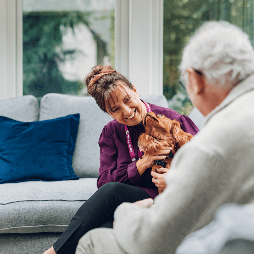 Person with short curly hair wearing a black sweater, standing against a pink background. - Home Instead
