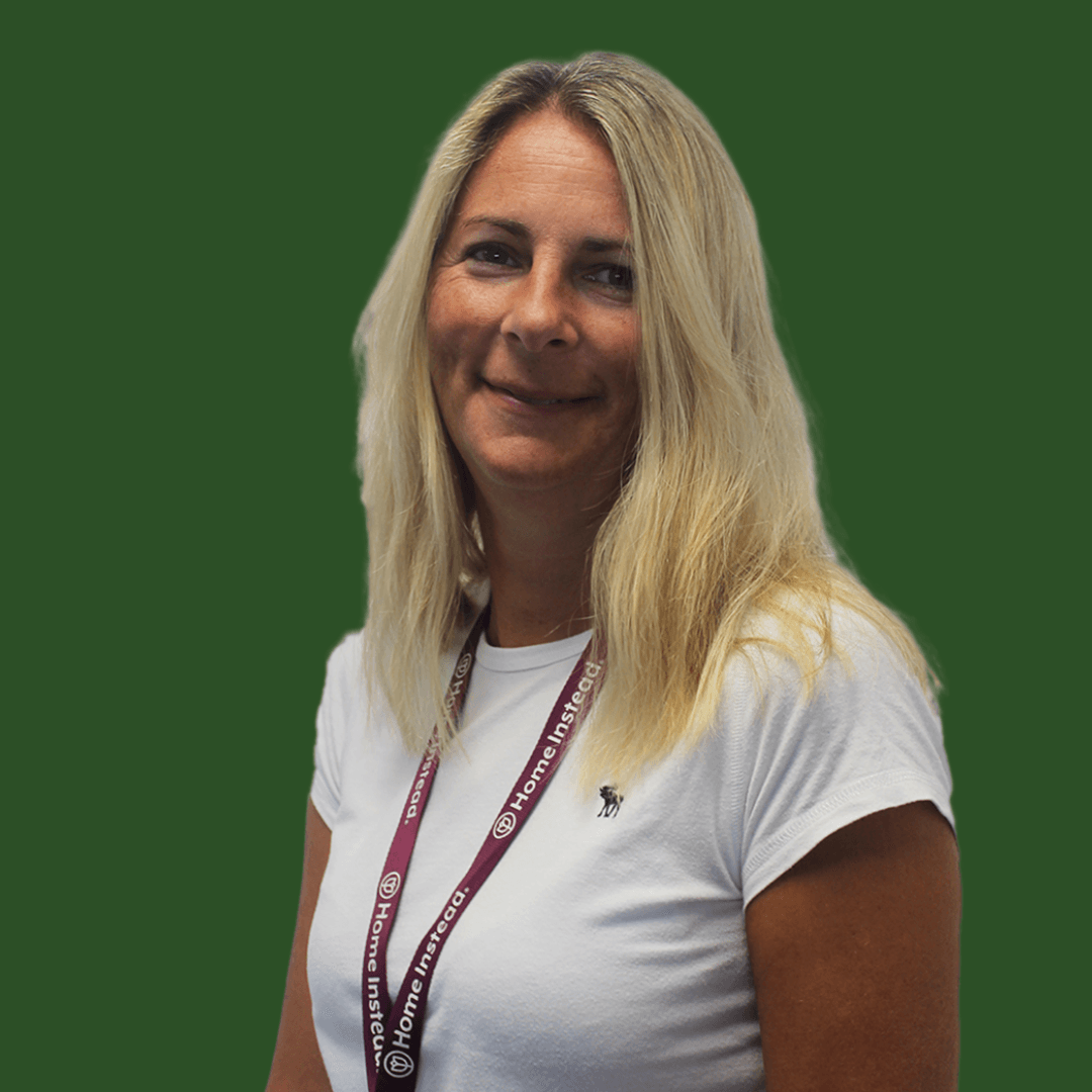 A woman smiles while kneading dough on a counter; an elderly man sits nearby in a sunlit kitchen. - Home Instead