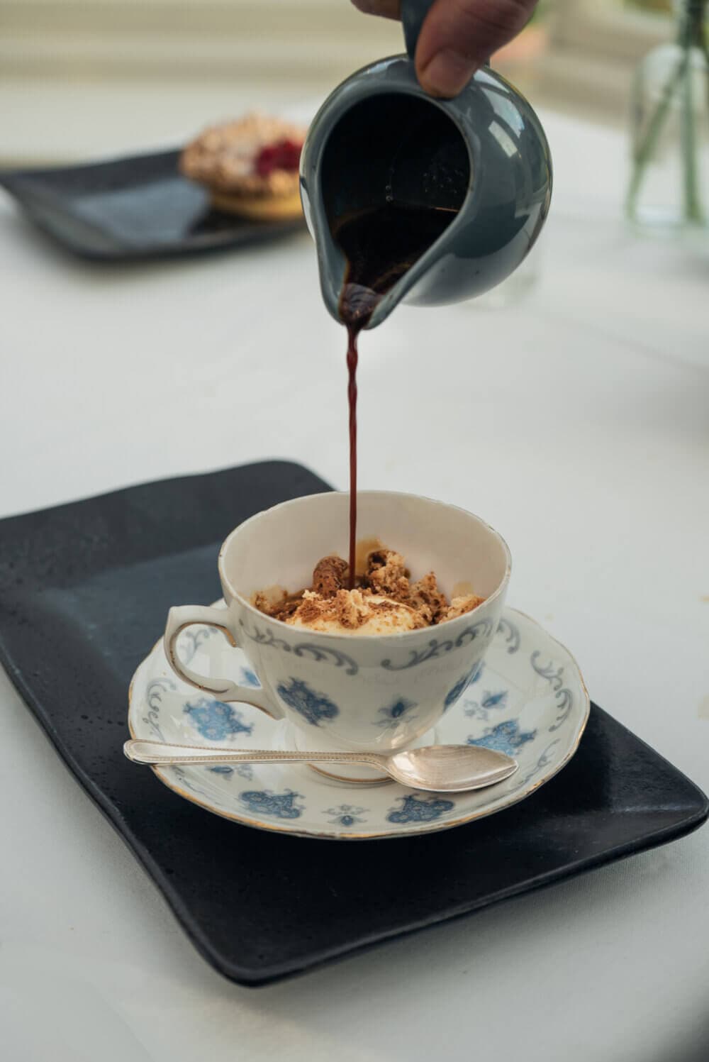 A person pours liquid from a jug into a decorated teacup with dessert on a matching saucer and spoon. - Home Instead