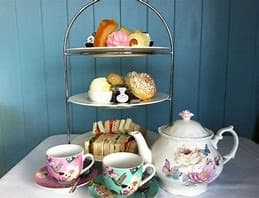 A tiered serving tray with assorted pastries, sandwiches, a teapot, and two teacups on a table against a blue wall. - Home Instead