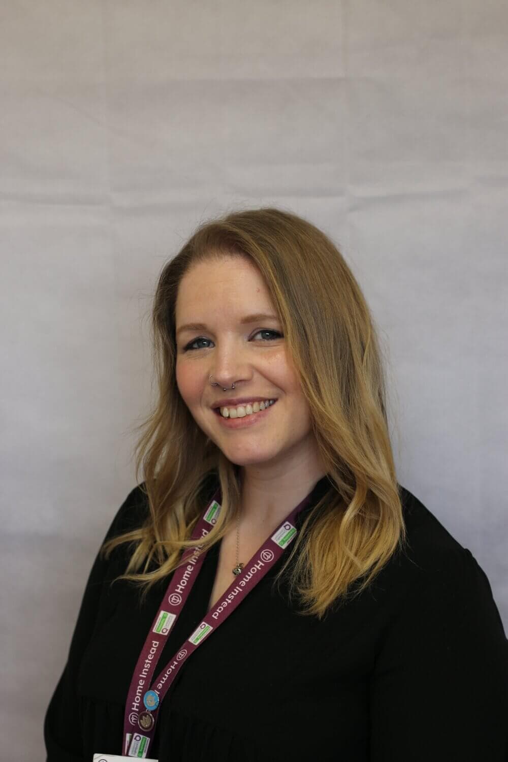 A woman with blonde hair and a black top smiles, wearing a lanyard with multiple tags, standing against a plain background. - Home Instead
