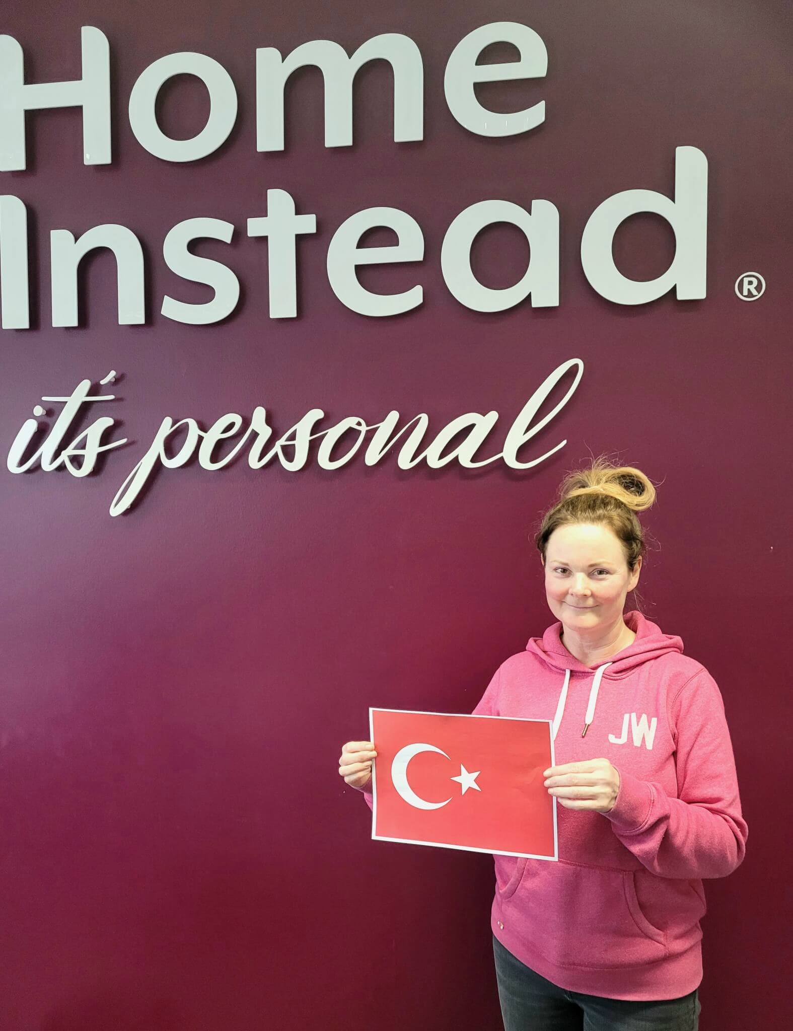 A person in a pink hoodie holding a Turkish flag, standing in front of a sign that says "Home Instead, it's personal. - Home Instead