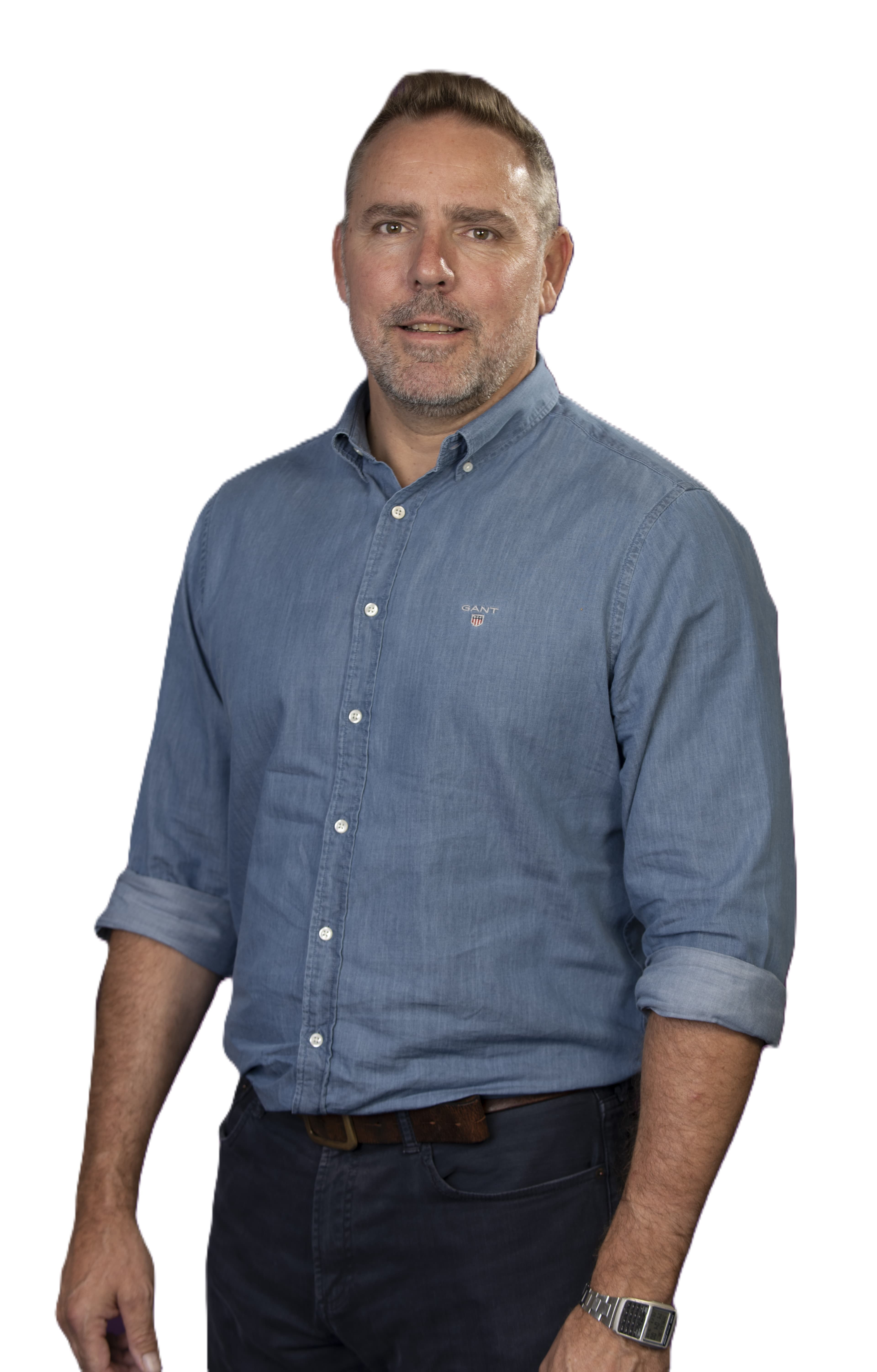 Man in a blue button-up shirt and dark pants standing against a white background, looking at the camera. - Home Instead