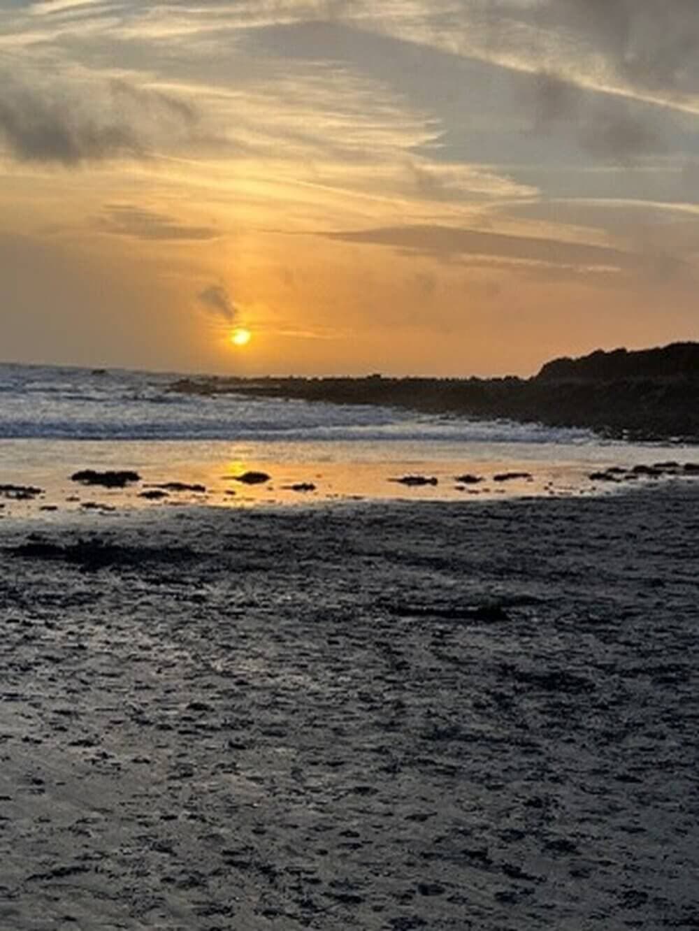 Sunset over a rocky shoreline with a mix of clouds and clear sky, reflecting on the wet sand and water. - Home Instead