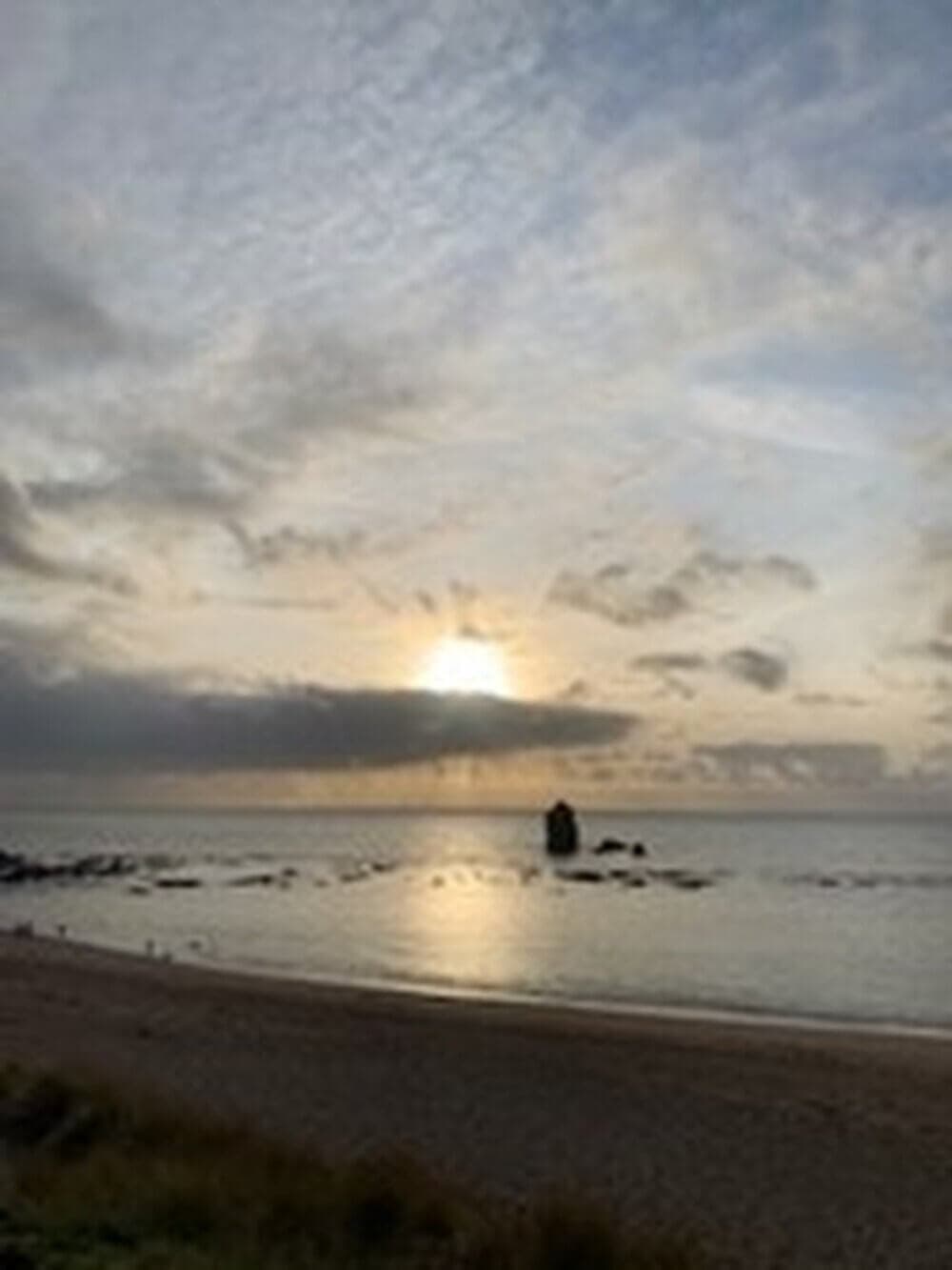 Sunset over a beach with gentle waves and a rock formation in the water, beneath a partly cloudy sky. - Home Instead