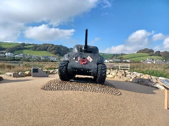 Front view of a black tank on display outdoors with a wreath on the front, green hills and houses in the background. - Home Instead