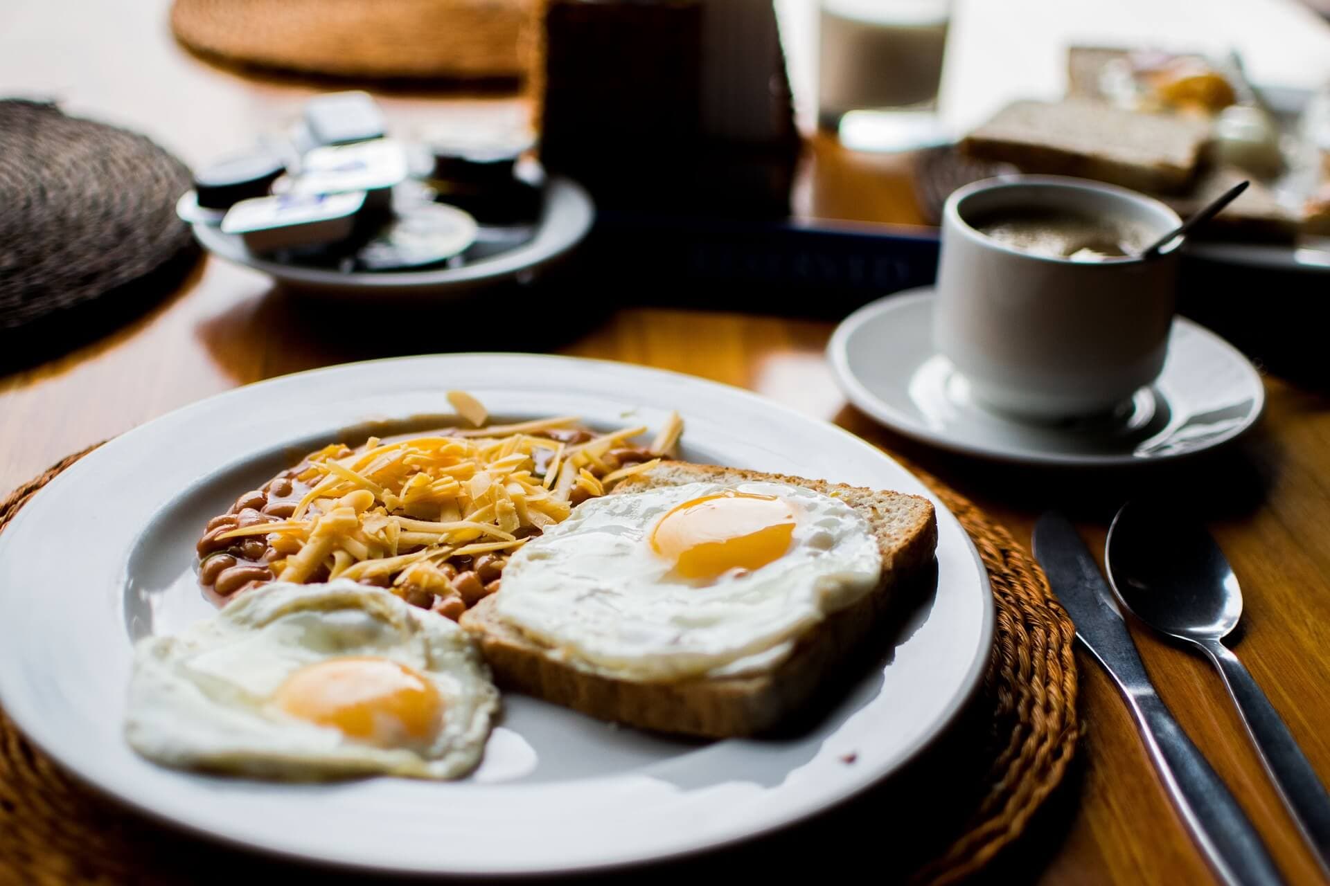 A breakfast plate with sunny-side-up eggs, toast, beans topped with cheese, beside a cup of coffee on a table. - Home Instead
