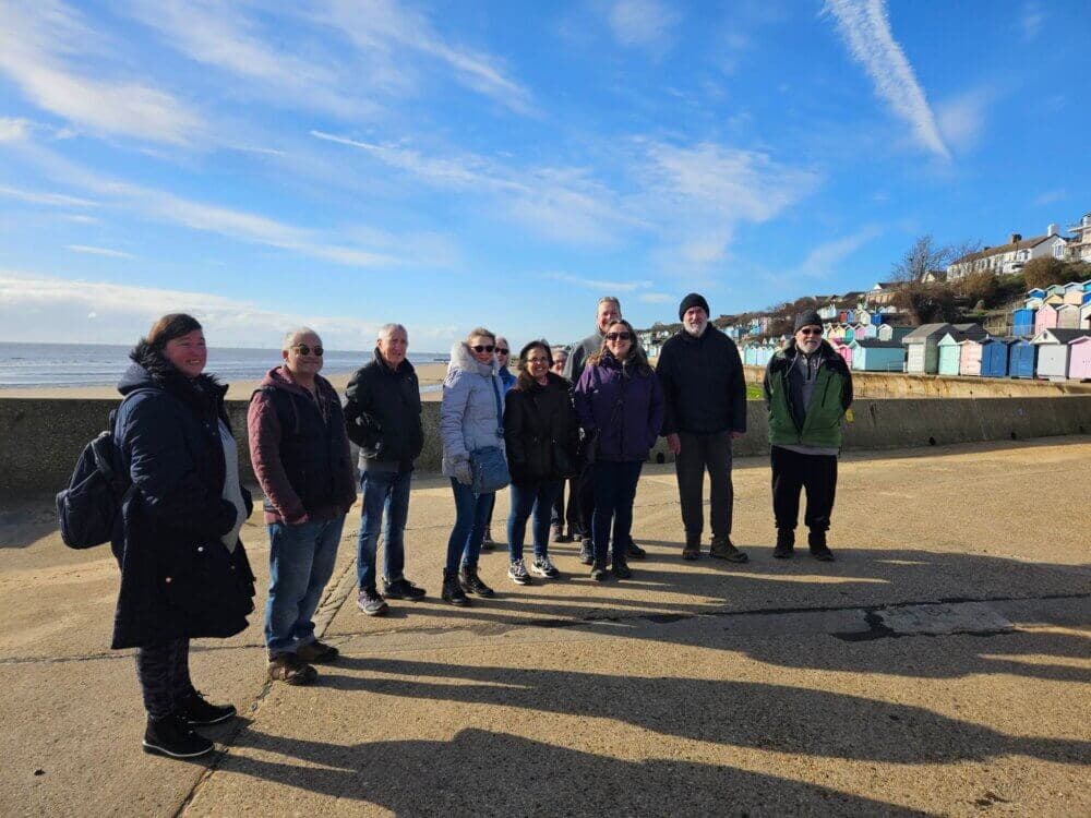 A group of people standing on a sunny beach promenade with colorful beach huts in the background. - Home Instead