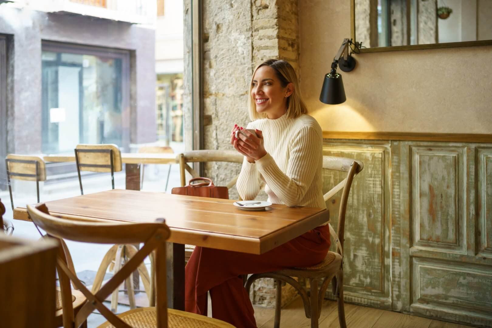 A woman in a cozy cafe, sitting at a wooden table, smiling, and holding a cup of coffee. - Home Instead