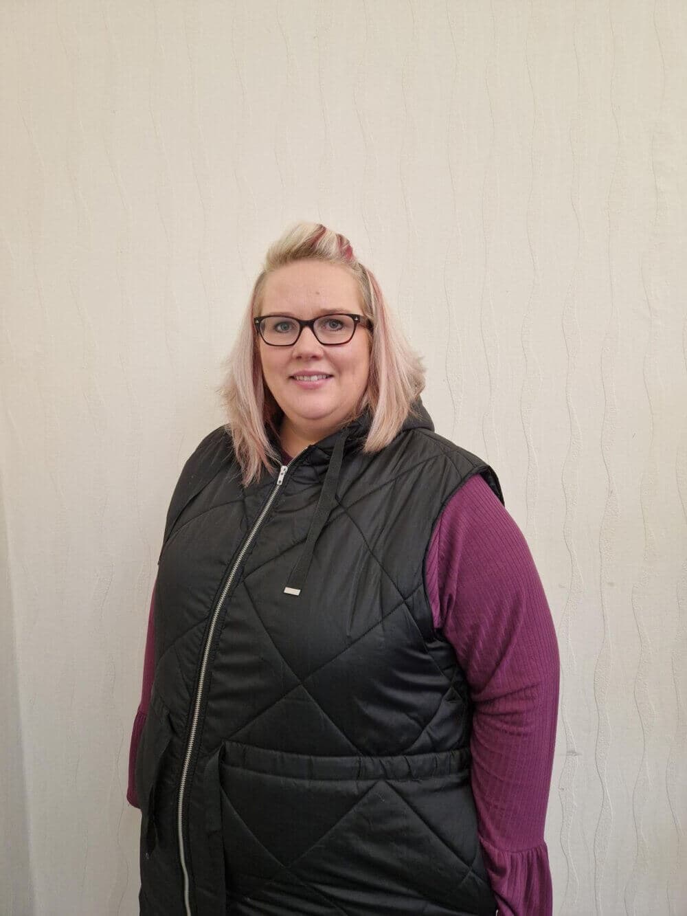 Person with blond hair wearing glasses, a black vest, and a purple long-sleeve shirt, standing against a textured wall. - Home Instead