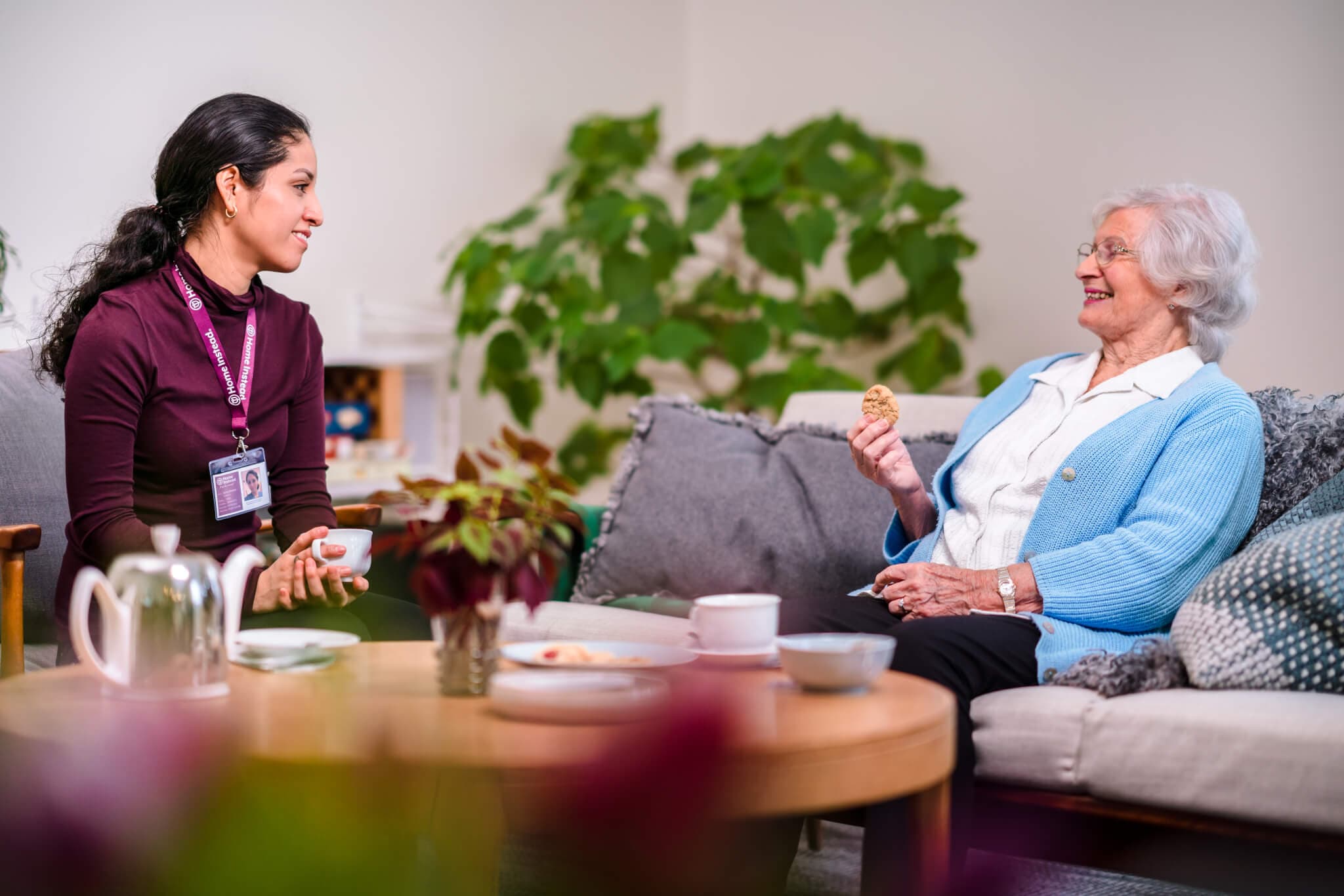 A caregiver and an elderly woman sit on a couch, smiling and enjoying tea and cookies together in a cozy living room. - Home Instead