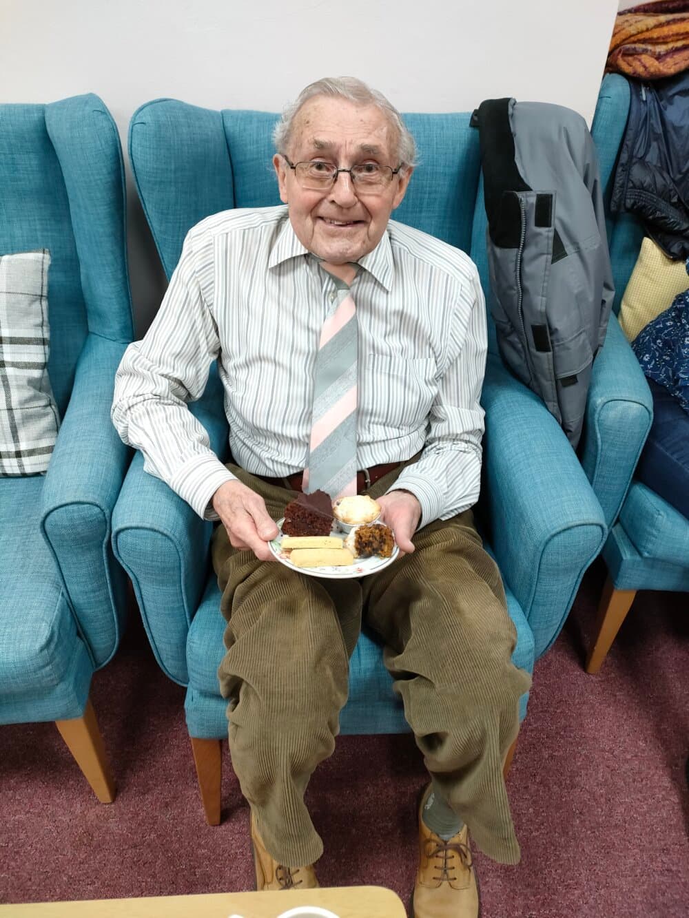 Elderly man in glasses and striped shirt sits on a blue chair, holding a plate of desserts, smiling at the camera. - Home Instead
