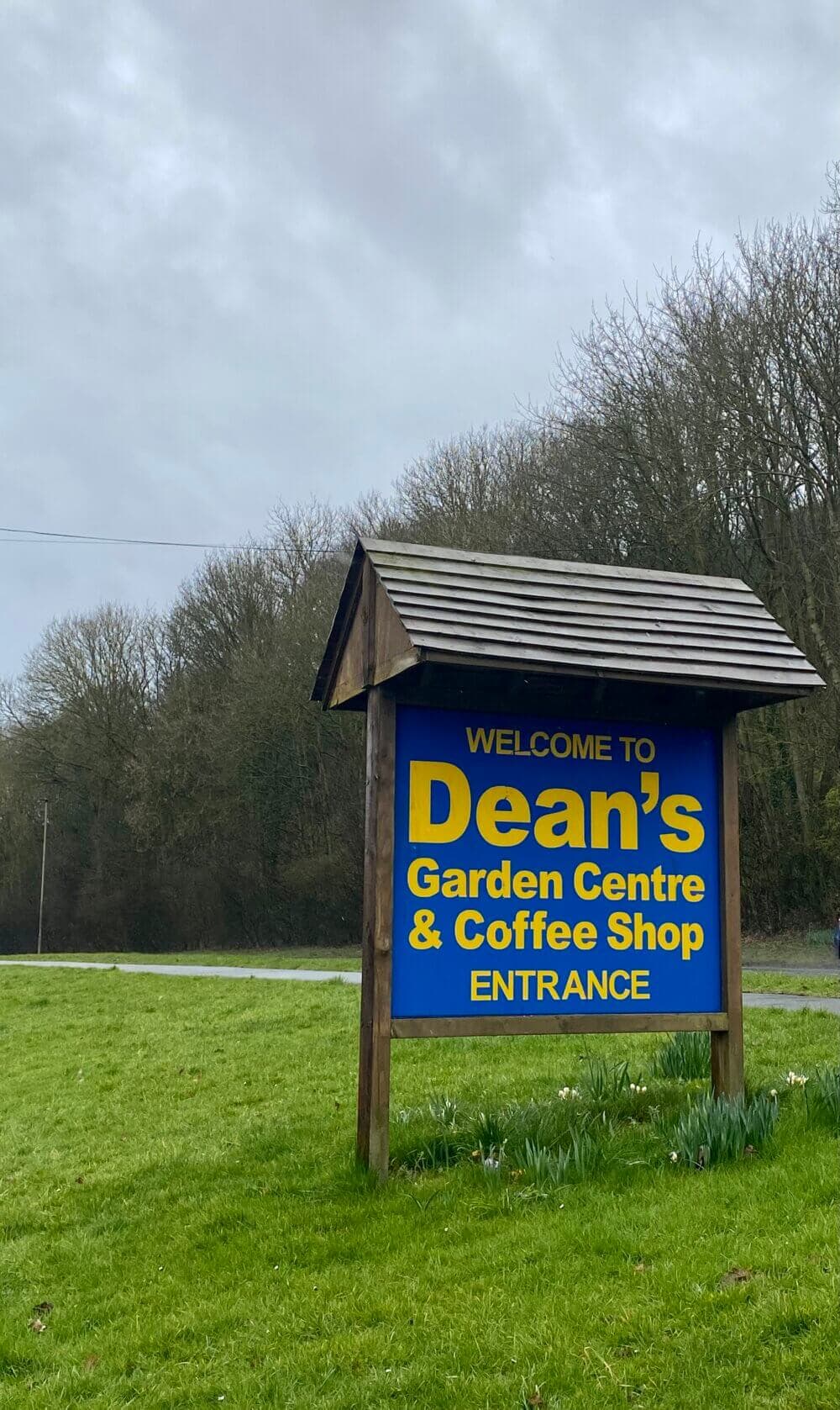 Sign reading "Welcome to Dean's Garden Centre & Coffee Shop ENTRANCE" on a lawn, trees in the background. - Home Instead