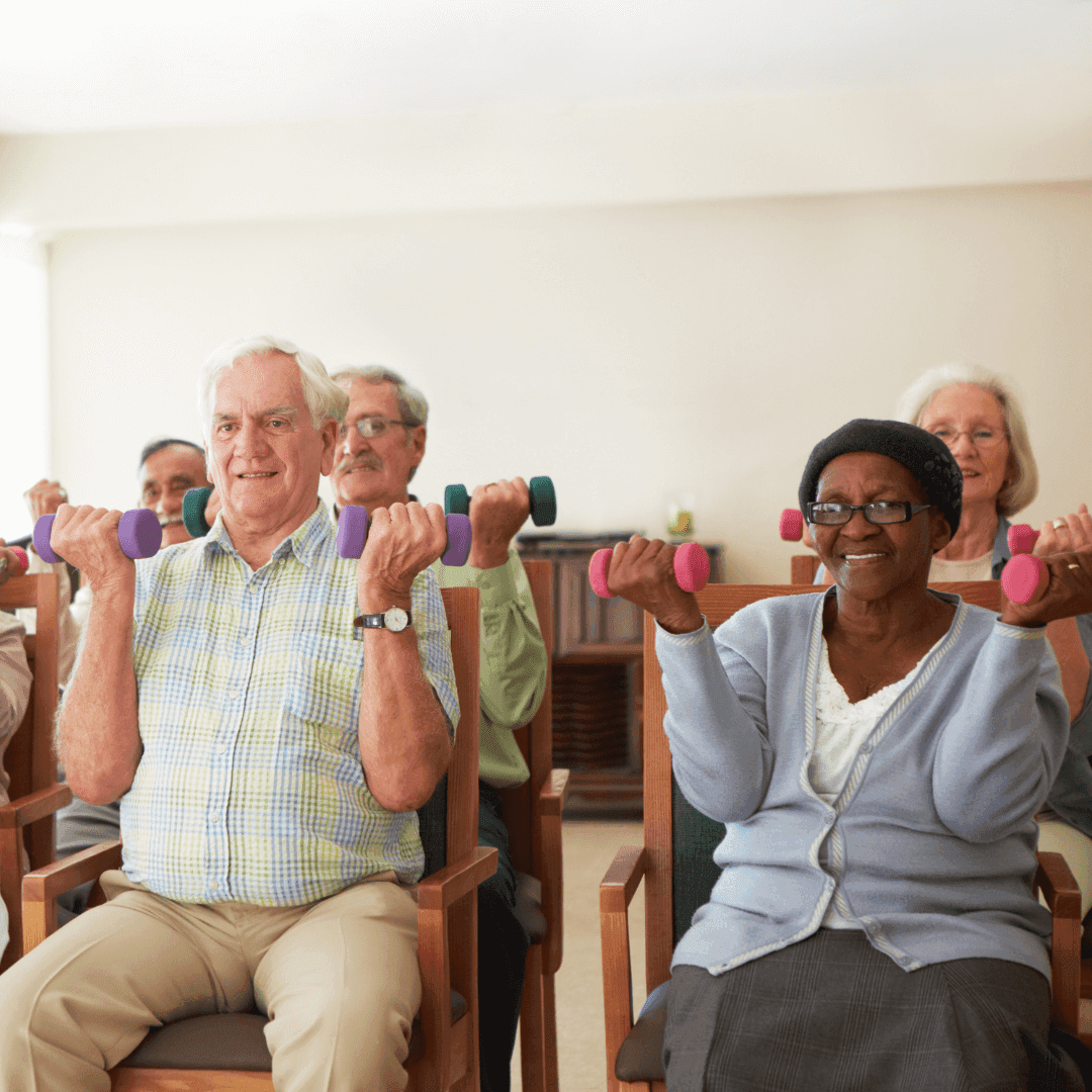 Seniors sitting on chairs, lifting dumbbells while smiling during an exercise class in a bright room. - Home Instead