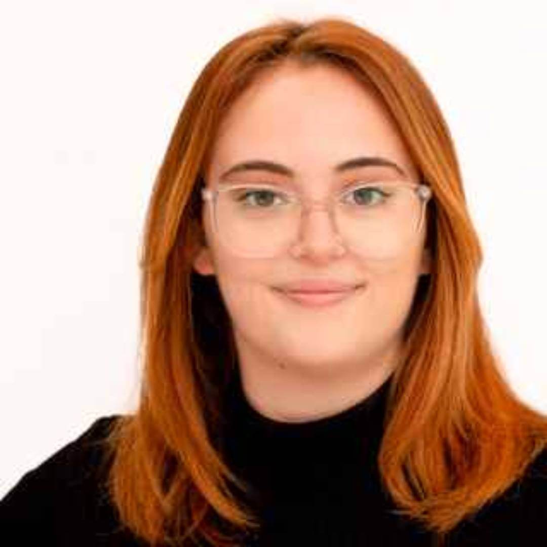 A woman with red hair and glasses smiles at the camera, wearing a black top against a plain white background. - Home Instead