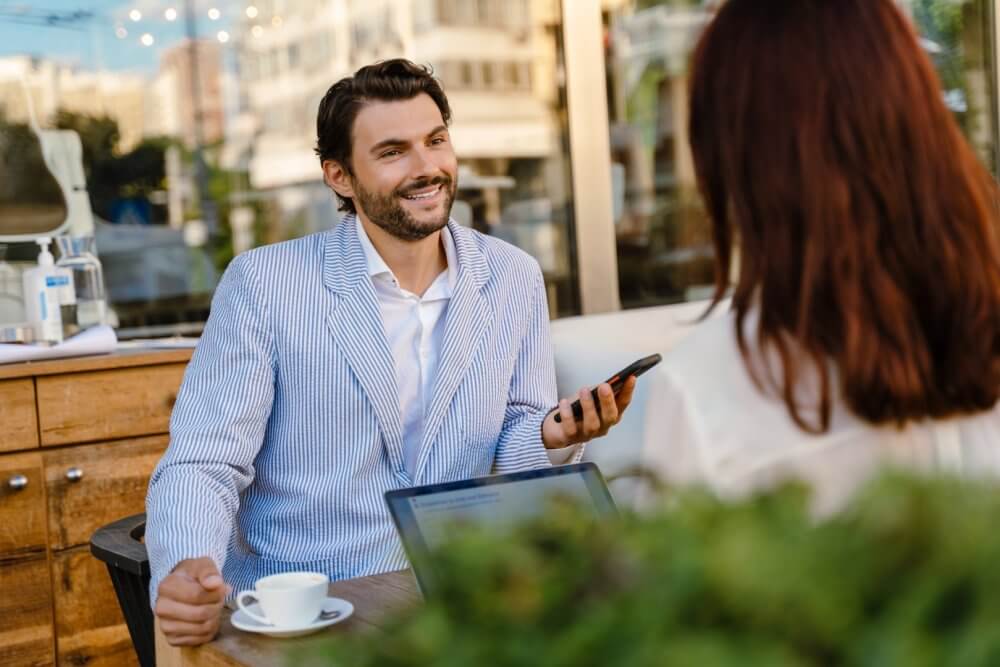 A man in a striped blazer smiles while holding a phone, sitting at a cafe table with a woman and a laptop. - Home Instead