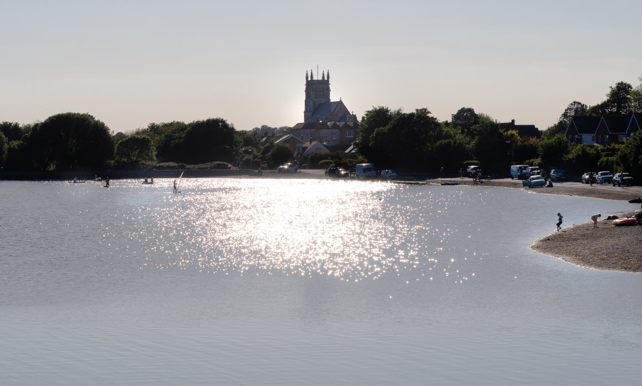 A church tower silhouetted against the setting sun, with shimmering water and people along the shore in the foreground. - Home Instead