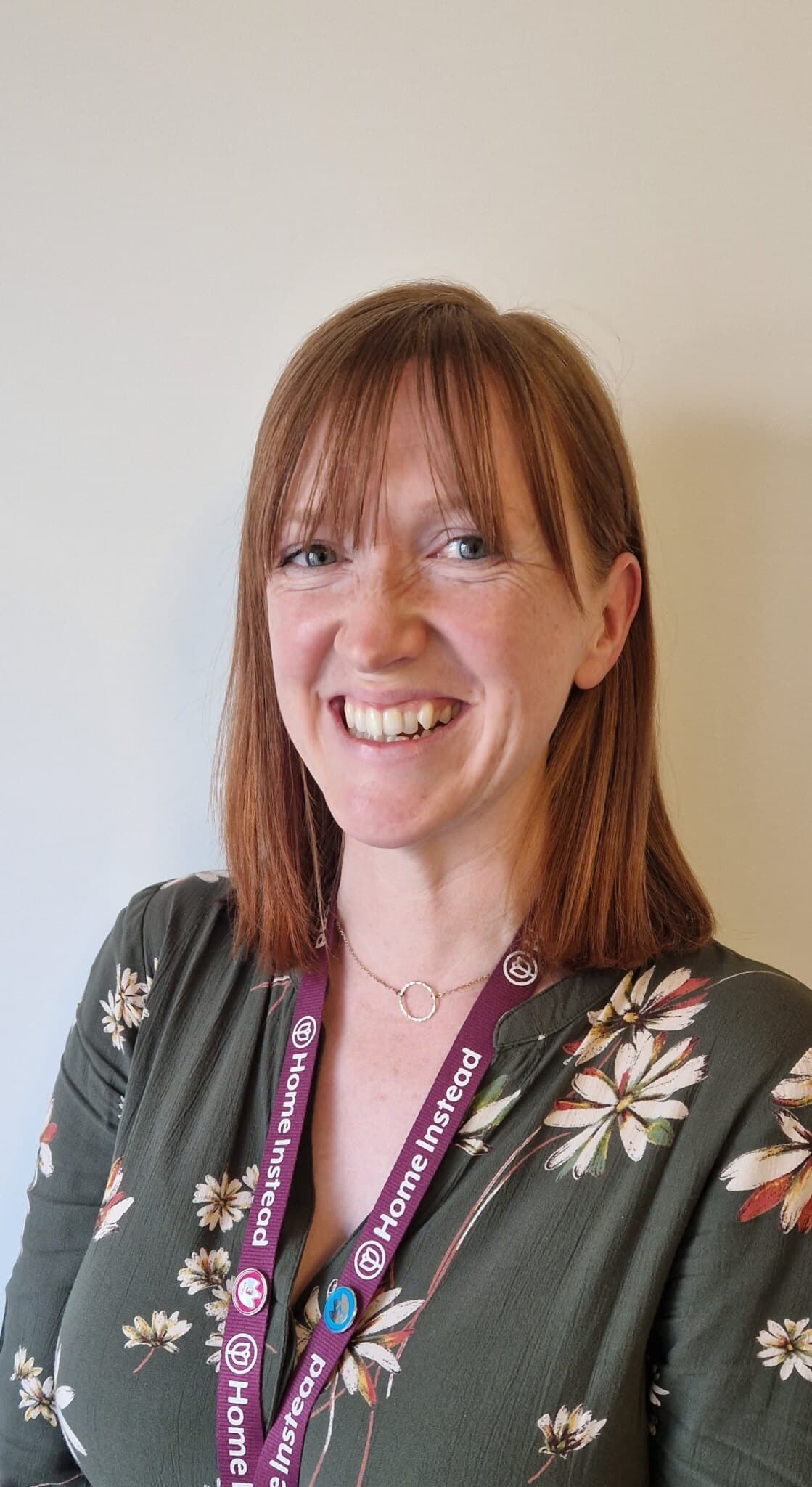 Smiling woman with shoulder-length hair, wearing a floral blouse and lanyard, standing against a plain background. - Home Instead