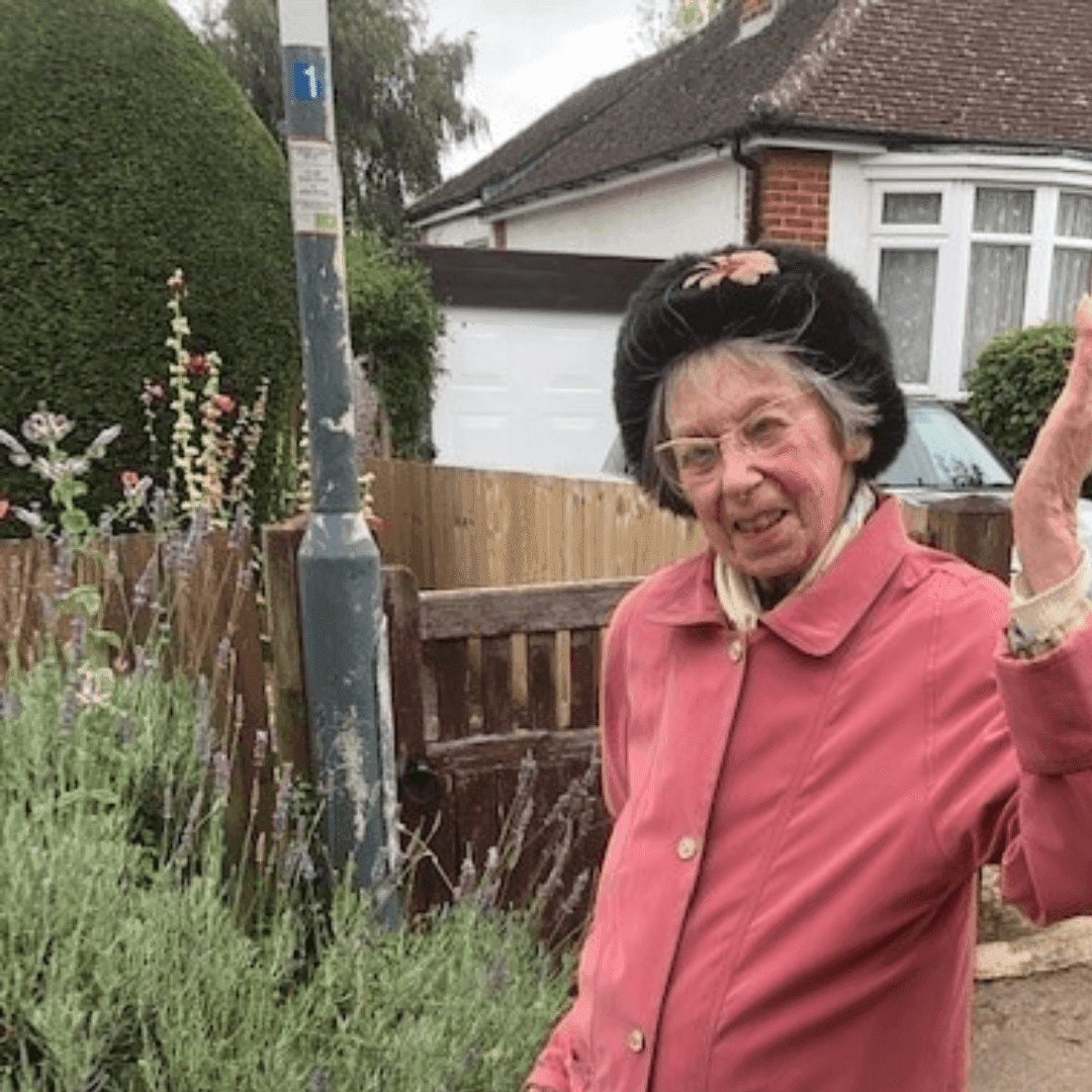 Elderly woman in a pink coat and black hat raising her hand while standing in front of a suburban house. - Home Instead