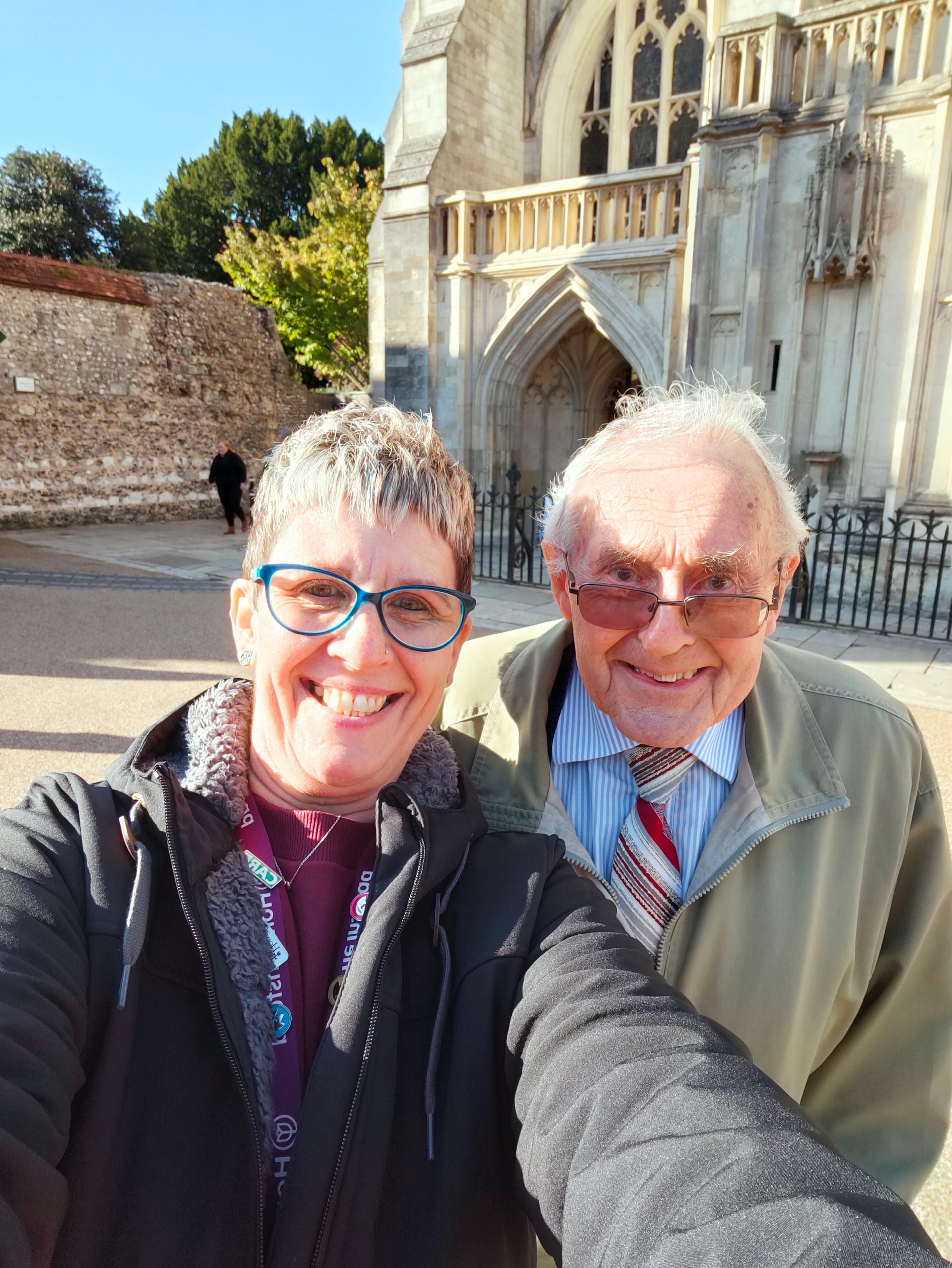 Two people smiling while posing for a selfie outside a historic stone building with an arched entrance. - Home Instead