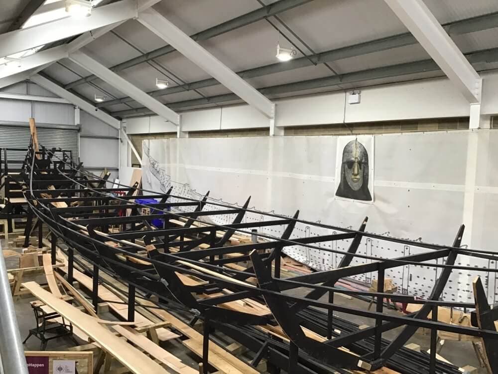 A large wooden boat frame under construction inside a spacious warehouse with a metal roof and walls. - Home Instead