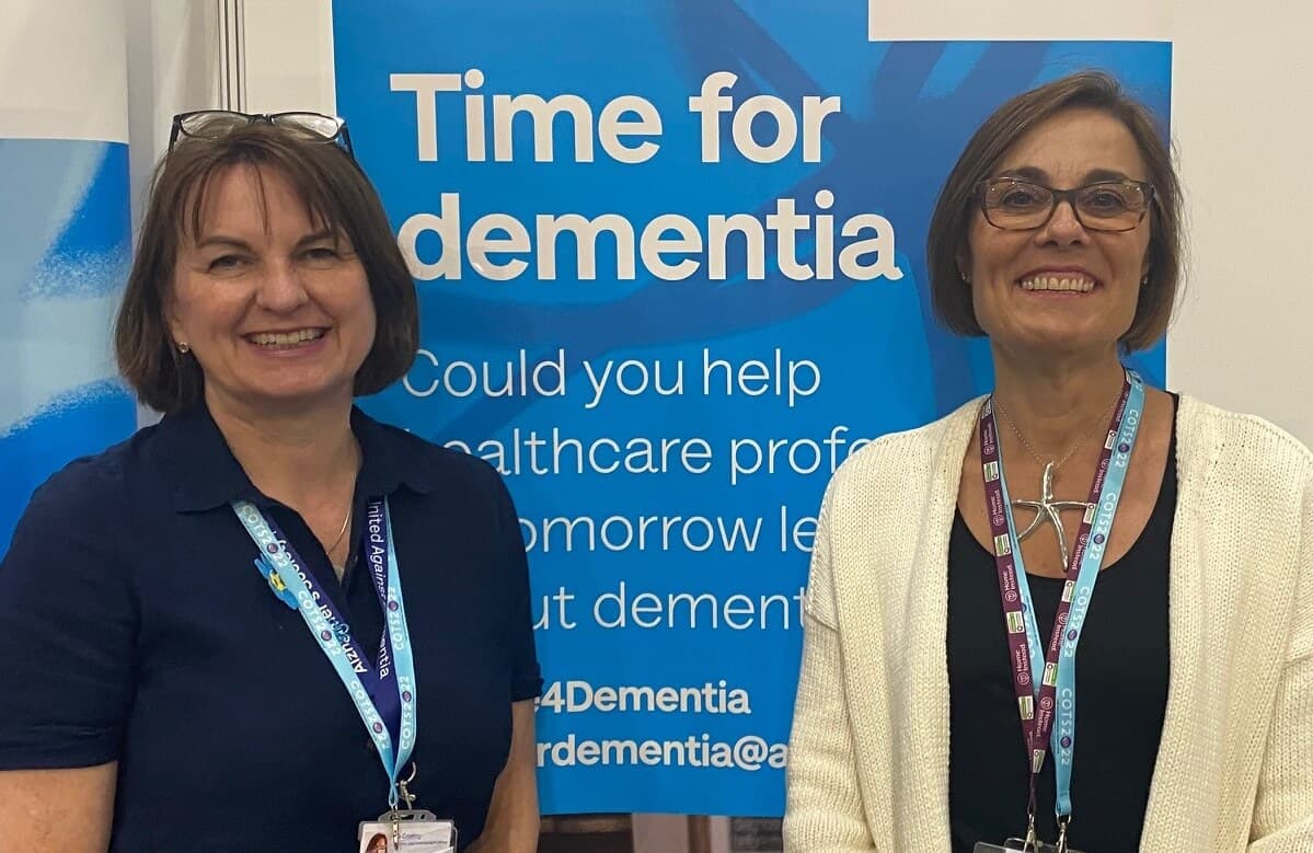 Two women smiling, standing in front of a sign that reads "Time for Dementia". - Home Instead