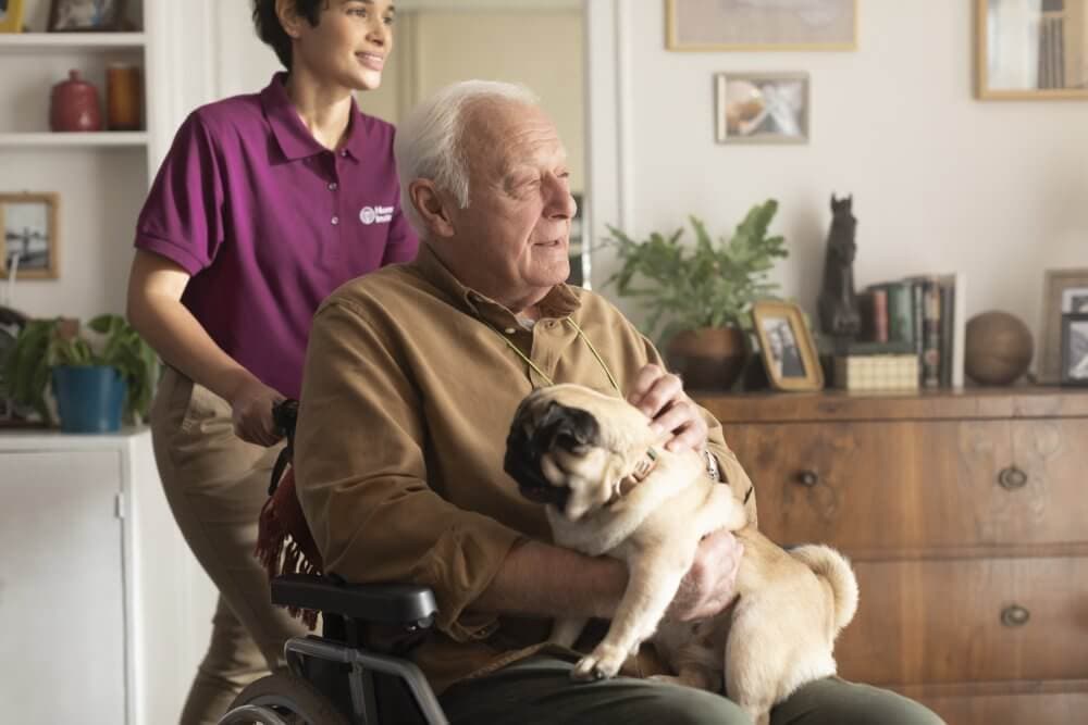 An elderly man in a wheelchair holding a pug, with a caregiver standing behind him in a cozy living room. - Home Instead