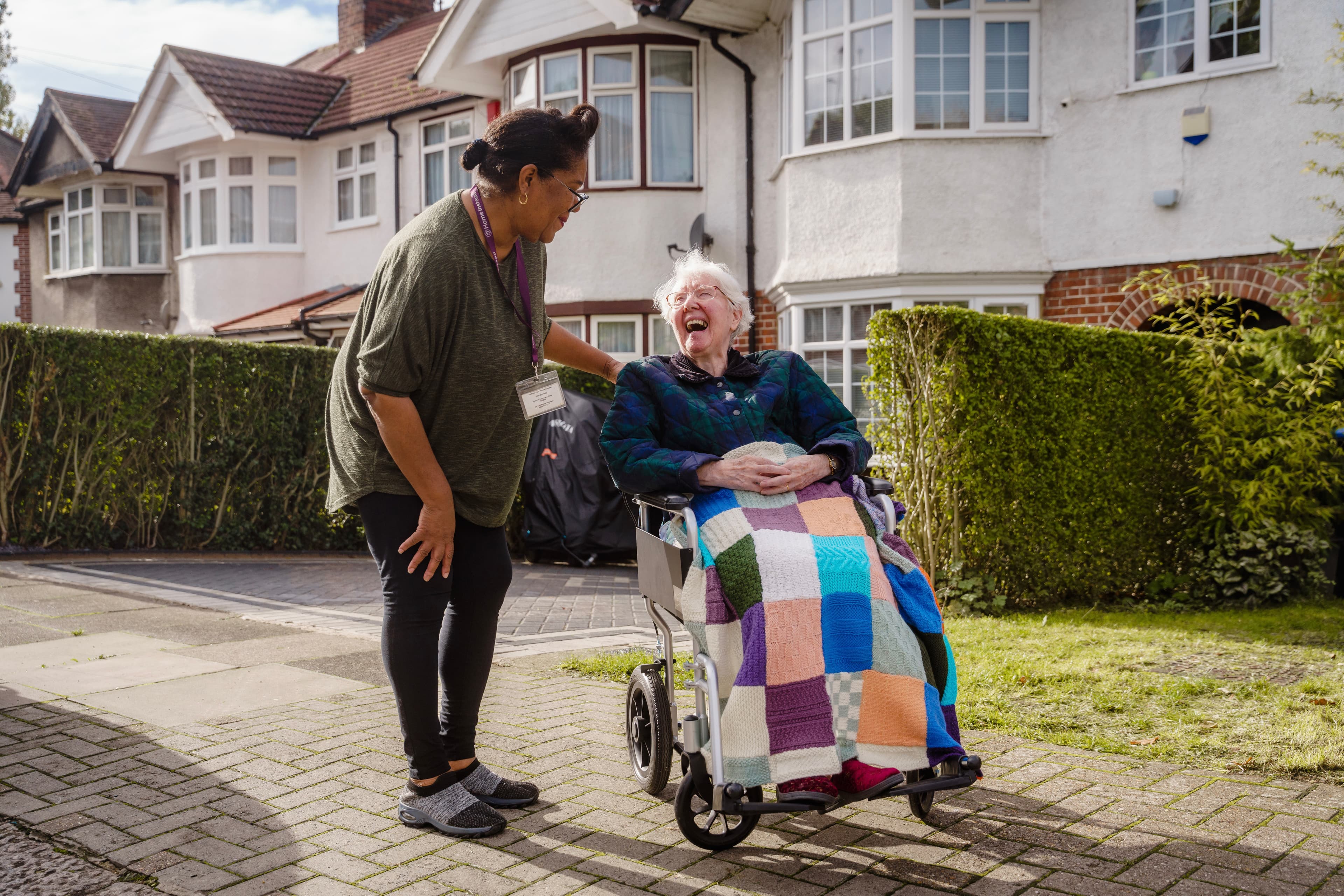 A caregiver and elderly woman in a wheelchair share a laugh outside in front of suburban houses on a sunny day. - Home Instead