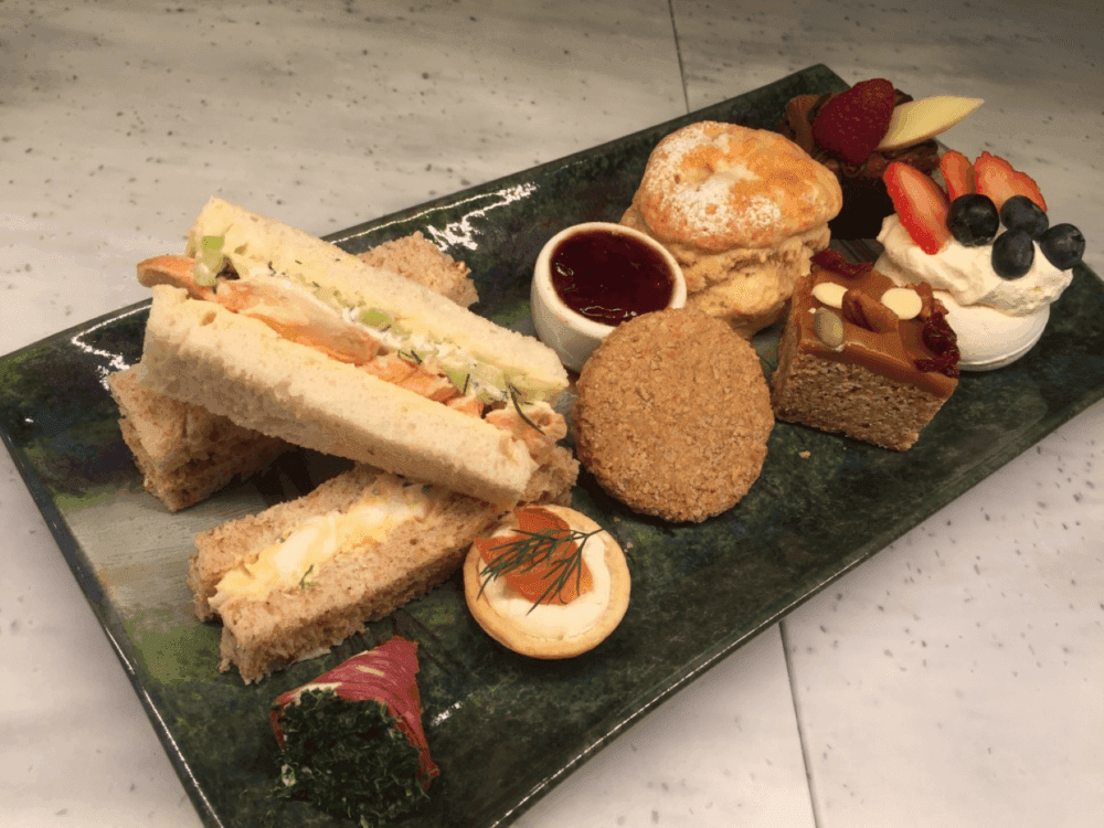 A plate with assorted sandwiches, a scone, a tart, a jam pot, and various pastries, garnished with fruits and herbs. - Home Instead