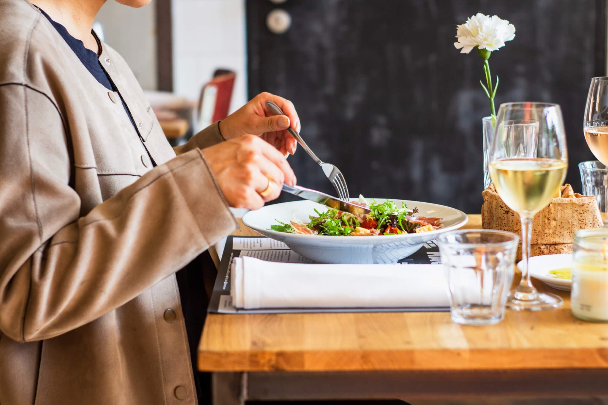 Person dining at a wooden table with a bowl of salad, wine glasses, and a flower vase. - Home Instead