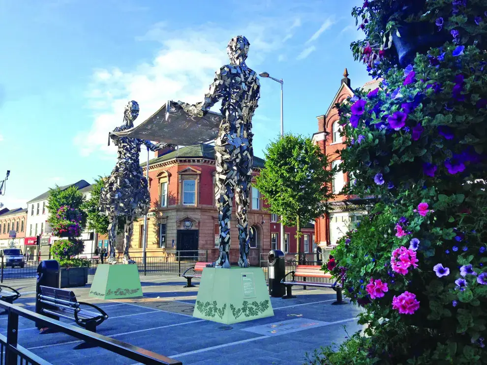 Two metal sculptures stand in a town square, surrounded by historic buildings and vibrant flowers. - Home Instead