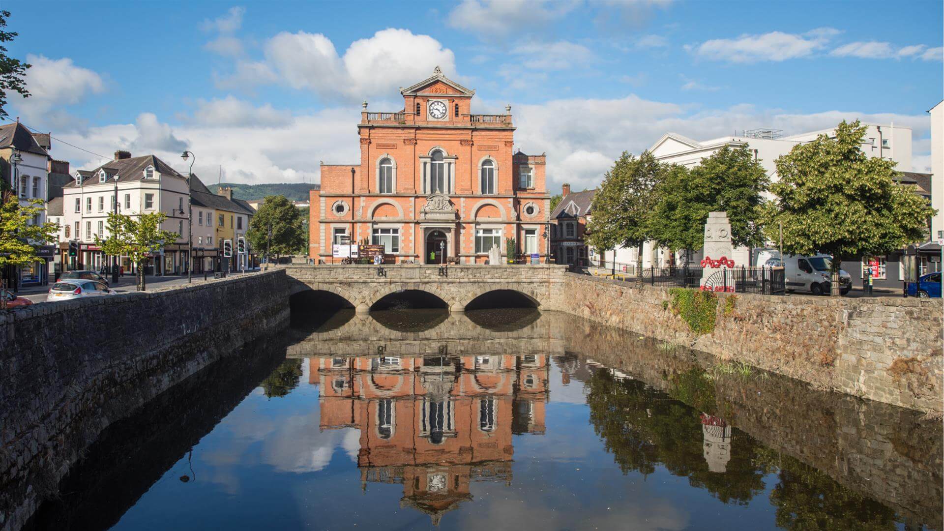A red-brick town hall building with a clock tower reflected in a river, surrounded by trees and cars. - Home Instead