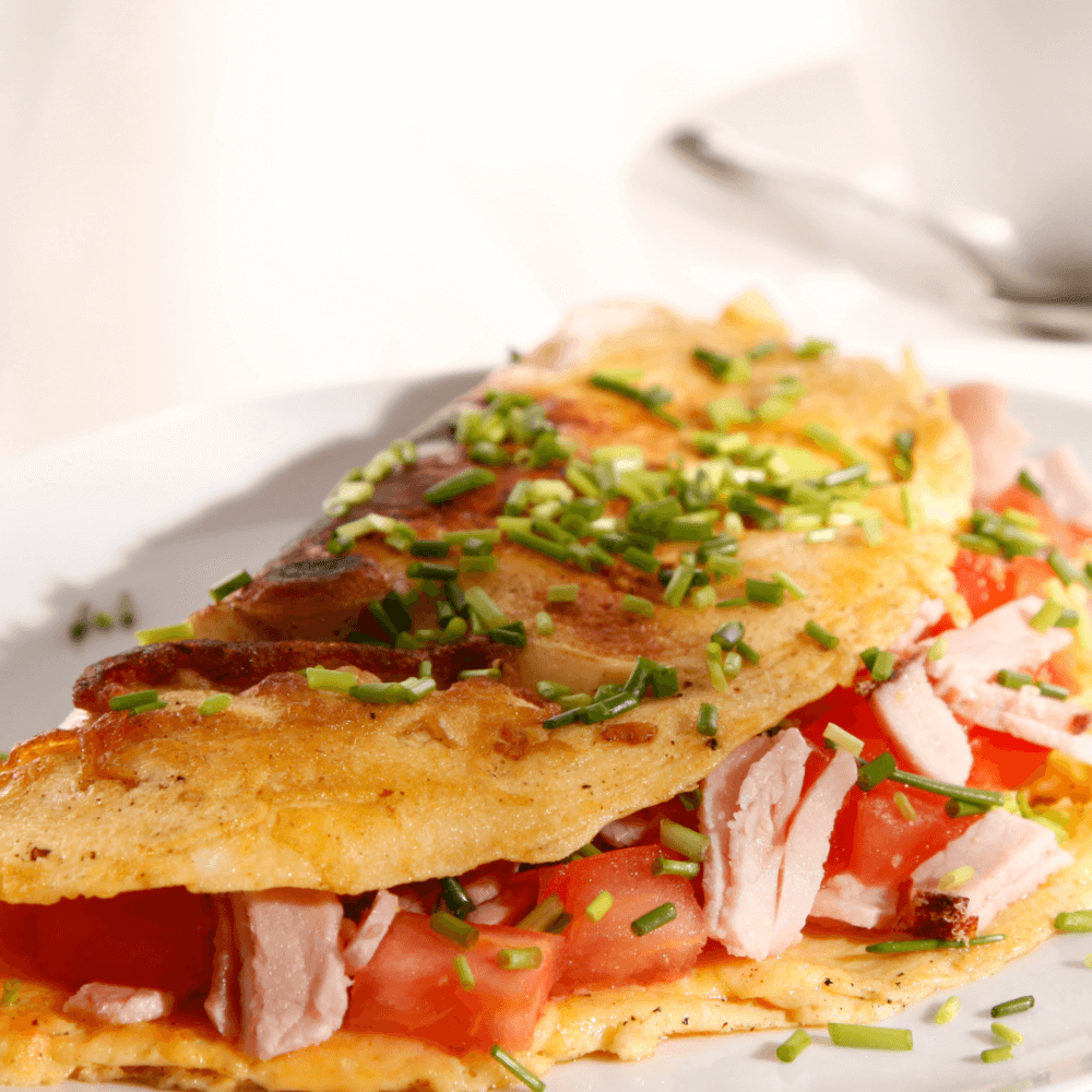 Close-up of a folded omelette filled with ham and tomatoes, garnished with chopped chives on a white plate. - Home Instead