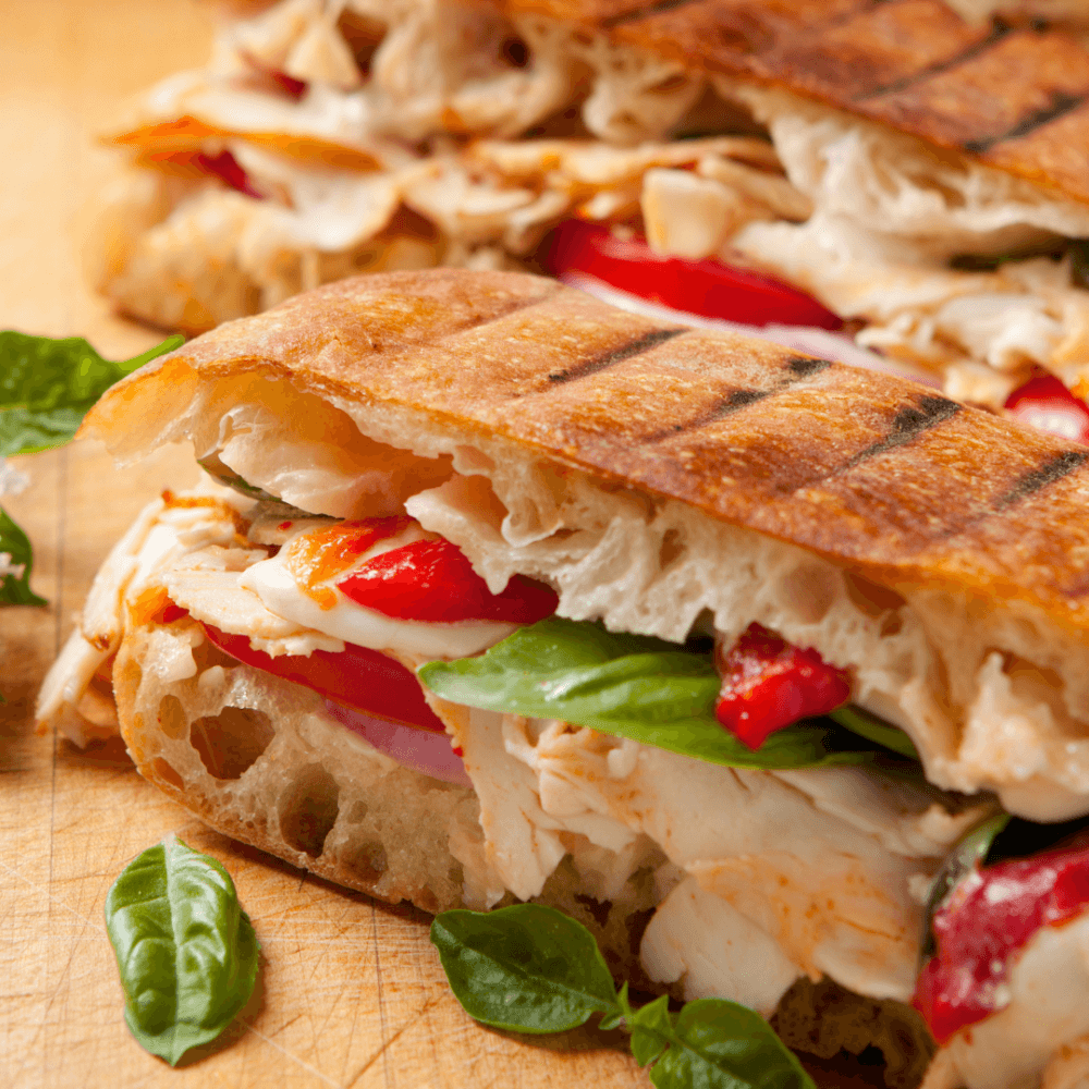 Close-up of a grilled chicken sandwich with tomatoes, cheese, basil, and roasted red peppers on a wooden surface. - Home Instead