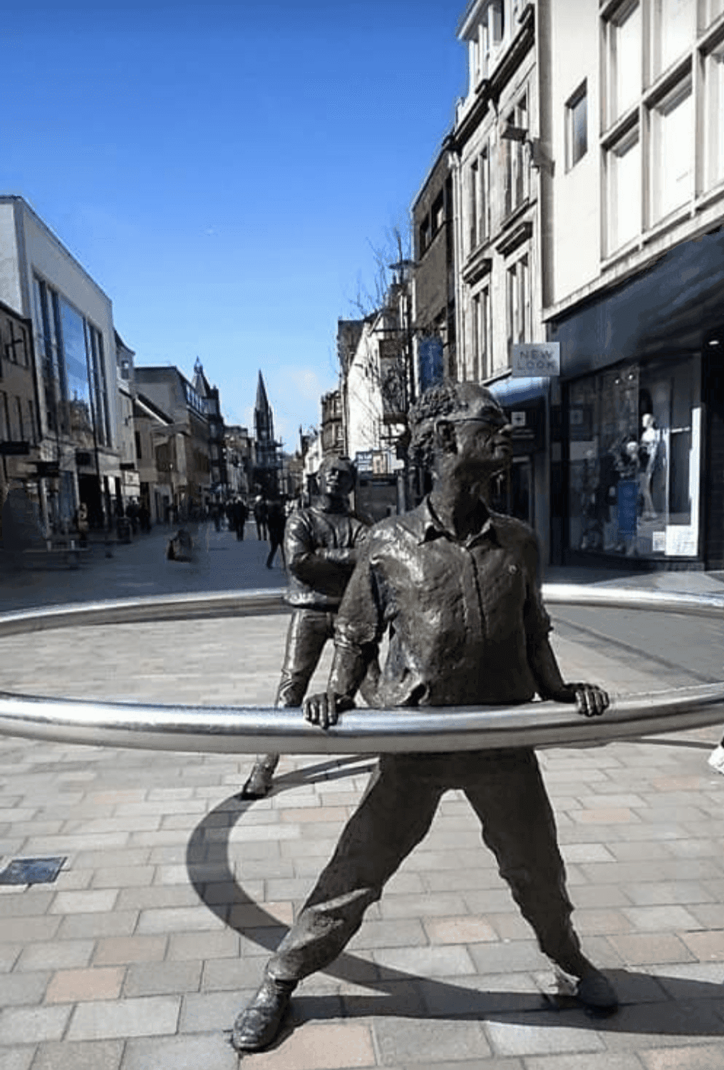 Sculptures of two men, each holding large hoops, on a pedestrian street lined with buildings and shops under a blue sky. - Home Instead