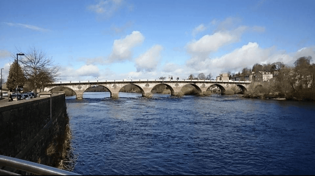 A stone bridge with multiple arches spans a wide, calm river under a blue sky with scattered clouds. - Home Instead